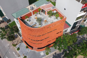 A brick building with a red roof and a rooftop patio featuring potted plants and chairs, set against a city view