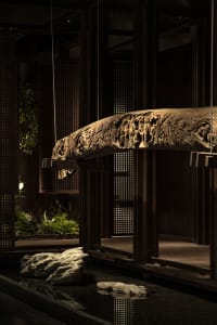 Intricate wooden structure with a large carved beam spanning the width of the image, accompanied by smaller wooden elements and a chair