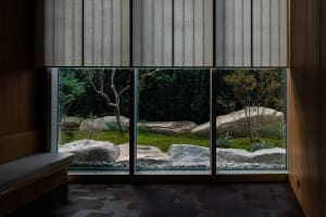 A cozy indoor space with a large window, rock wall, potted plants, and vases, offering a view of a garden outside