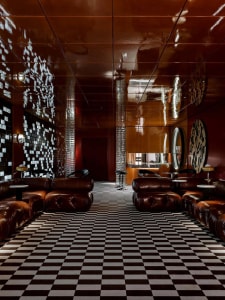 A modern living area with a checkered floor, leather chairs, and a mix of wood and metal materials, featuring warm lighting and a comfortable atmosphere