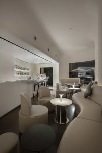 A modern restaurant bar with a counter and chairs, featuring white walls and flooring, and bottles on the counter