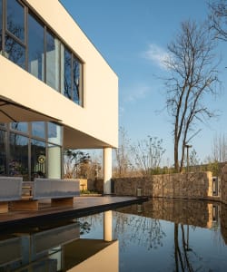 Modern house with a large glass wall overlooking a pond, featuring a living area with couches and chairs, and an exterior made of concrete