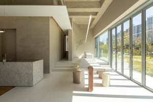 Open modern living area with natural light from a large window, featuring tables and benches in a minimalist design