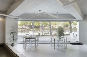 A modern restaurant interior with glass walls, multiple dining tables, and chairs, surrounded by natural light