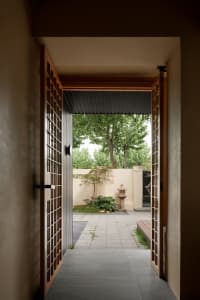 A serene outdoor entrance with a wooden-framed glass doorway, surrounded by two trees and a calm atmosphere