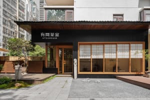 Modern Asian restaurant with a black facade, wooden elements, and natural light from windows, featuring a dining area with tables and chairs and potted plants