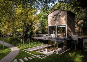 A house with a brick exterior and large windows surrounded by trees in a forest, featuring chairs and potted plants