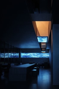 A modern restaurant with a large dining table, ocean wave screens on the walls, and a comfortable atmosphere