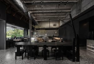 Empty modern restaurant interior with a large dining table, chairs, potted plants, concrete walls, and dim lighting