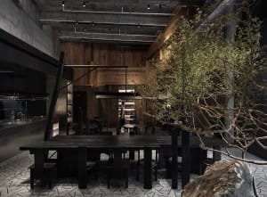 A cozy restaurant dining area with a large wooden table surrounded by black chairs, bottles, and an inviting atmosphere