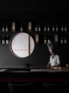 A professional chef in an Asian restaurant kitchen, surrounded by bottles and wine glasses, preparing food with focus and care