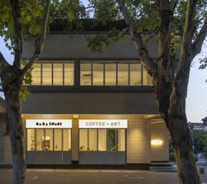A modern building with two storefronts, a coffee shop, and an art gallery, featuring large windows, potted plants, chairs, and dining tables
