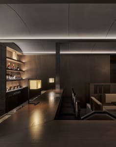 A modern bar area with a wooden dining table surrounded by chairs, bottles, and warm lighting