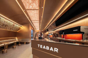 A well-lit cafe interior with a counter, bar stools, and TV screens, featuring a brown surface and comfortable seating arrangements