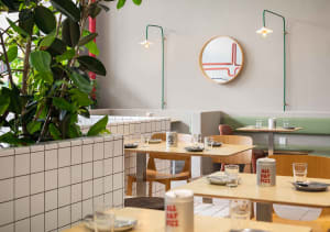 A cozy restaurant interior with wooden tables, chairs, and potted plants, set up for customers with cups and a welcoming atmosphere