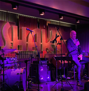 A restaurant interior with a stage, purple ambiance, chairs, couches, and microphones, designed for live performances or events
