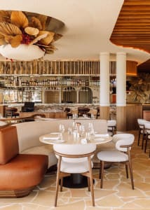 A modern restaurant interior with a dining area featuring wooden elements, tables, chairs, and a bar, with wine glasses and an inviting atmosphere