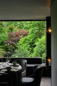 Dining room with a wooden table and chairs, surrounded by wine glasses and bowls, with a large window providing a view of trees outside