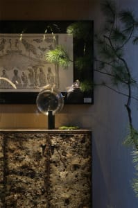 A living area with a stone table, glass bowl, and several potted plants, featuring three decorative figures