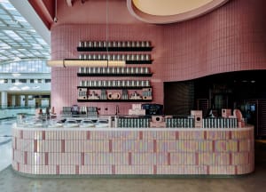A pastel pink cafe interior with a well-organized display counter featuring bottles and cups on multiple levels