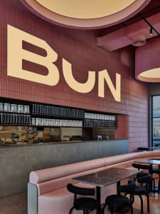A cozy cafe interior with a pink accent wall, dining tables, chairs, and a sign reading 'Bun'