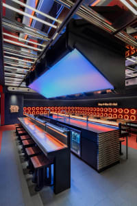 Industrial kitchen with a stainless steel island, black countertops, and multiple benches for seating, with a refrigerator and chairs scattered throughout