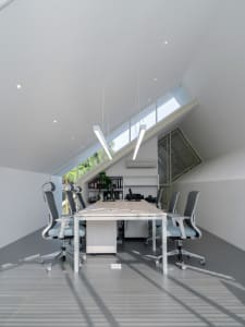 A well-lit office space with a vaulted ceiling, a dining table, chairs, and potted plants, creating a comfortable workspace
