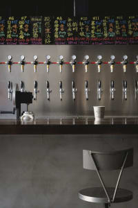 Industrial-style bar area with a long counter featuring 14 beer taps and two cups, set in a busy restaurant or bar atmosphere