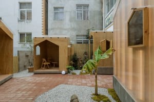 A serene outdoor space with two wooden structures, a garden shed and a small house/storage unit, surrounded by plants and trees, with chairs for relaxation