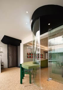 A modern living space with a green table, chairs, potted plants, and vases, surrounded by a large glass door and natural light