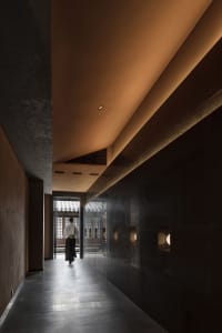 A woman in traditional clothing walks down a hallway with a stone wall, accompanied by two smaller figures in the background