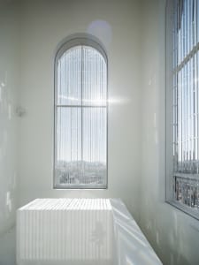 An empty white room with two beds and a chair, lit by natural light from a large window