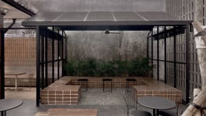 A brick-floored patio with stone benches and a metal table, shaded by a large umbrella, features a comfortable outdoor seating arrangement with three chairs around a dining table