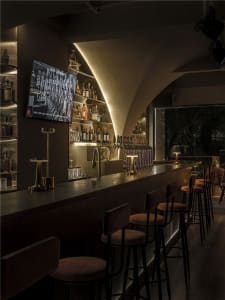 A bar area in a restaurant with a TV above a counter, bottles behind it, and comfortable seating for patrons