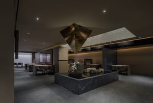 Dimly lit restaurant interior with a stone sculpture centerpiece, surrounded by tables and chairs, with a warm and earthy atmosphere