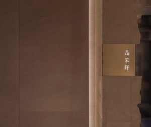 A modern building lobby with an elevator situated between two walls, one of which is a marble wall, and a gold sign above the elevator