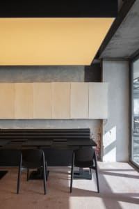 A modern cafe with a concrete wall, large windows, and a cozy atmosphere with three black chairs around a dining table