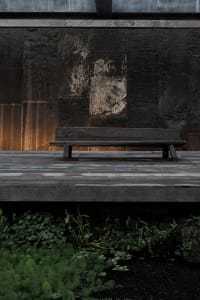 A serene outdoor patio area with a wooden bench on a stone platform surrounded by greenery