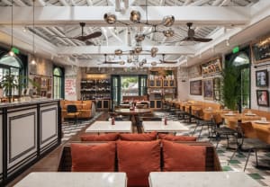 A restaurant interior with multiple tables, couches, and paintings, illuminated by hanging lights, creating a warm and inviting atmosphere