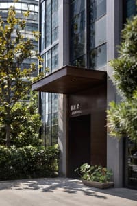 A simple Asian building with a brown doorway, potted plants, and a window above, set against a concrete background