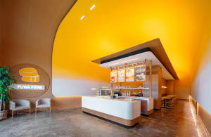 A modern Japanese restaurant interior with a yellow theme, featuring a counter with a cash register and displayed food items, surrounded by cups and a bowl with a spoon