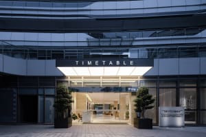 A modern office lobby with a large glass door, potted plants, chairs, and a clock, set against a well-lit background