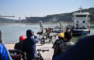 A bustling waterfront scene with people walking across a bridge between two boats, one larger than the other, with individuals taking photos and moving between the boats and shore