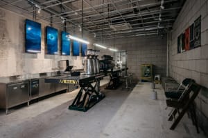 Industrial kitchen with concrete floors and walls, featuring a countertop with various items and chairs, lit by overhead lighting