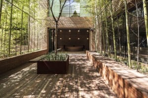 A serene outdoor space with a wooden deck, brick wall, and benches, surrounded by trees and natural light