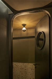 A dimly lit hallway with a black door, a round window above the doorway, and two light fixtures, one near the center and one to the right