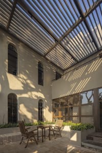 A modern outdoor patio with a dining table, chairs, potted plants, and benches, set against a clean-lined house with windows and doors