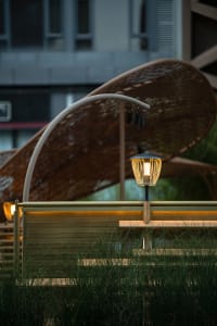 A wooden bench under a large umbrella in front of a building, with two lights providing illumination and a relaxing atmosphere