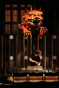 A professional chef prepares food in an Asian restaurant kitchen with a dragon head statue, surrounded by bowls and bottles, in a focused and professional atmosphere