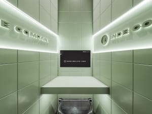 Bathroom with green tiles, two sinks, and a TV above one sink, in a well-lit space